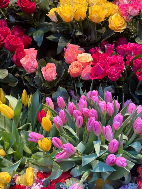 A vibrant display of fresh flowers, including pink and yellow tulips with elongated green leaves, along with clusters of pink, yellow, and peach roses arranged in a lush, densely packed bouquet. The f