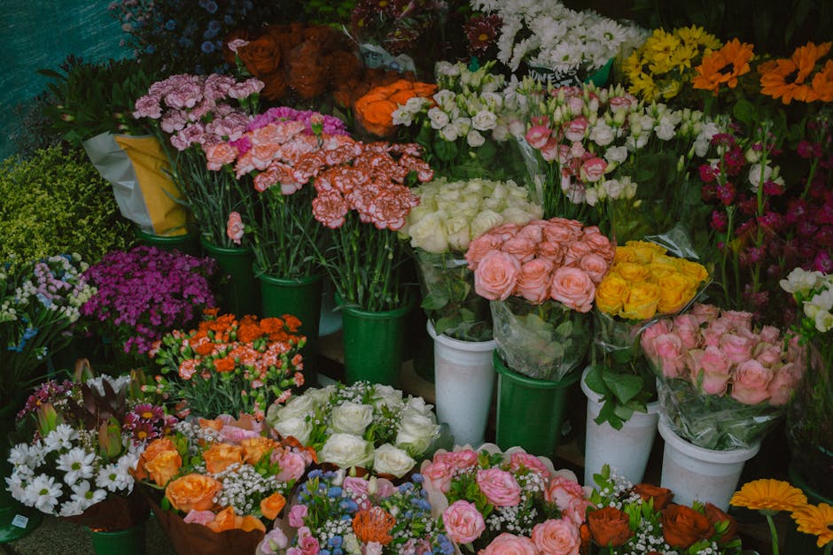 A vibrant collection of fresh flower bouquets and potted plants displayed in a florist shop, featuring roses, carnations, daisies, and other blooms in shades of pink, peach, white, purple, yellow, and